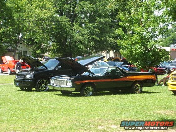 Black Ford Ranchero with orange stripe, hood up, parked on grass next to a black pickup truck